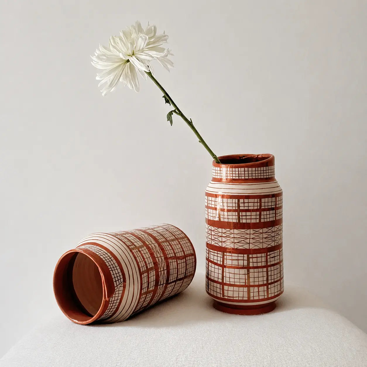 Two patterned ceramic vases in a terracotta and cream color scheme, one standing upright holding a white daisy and the other lying on its side, set against a neutral light gray background.