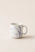 A white ceramic coffee mug with a blue abstract sketch design on the side, set against a solid beige background.