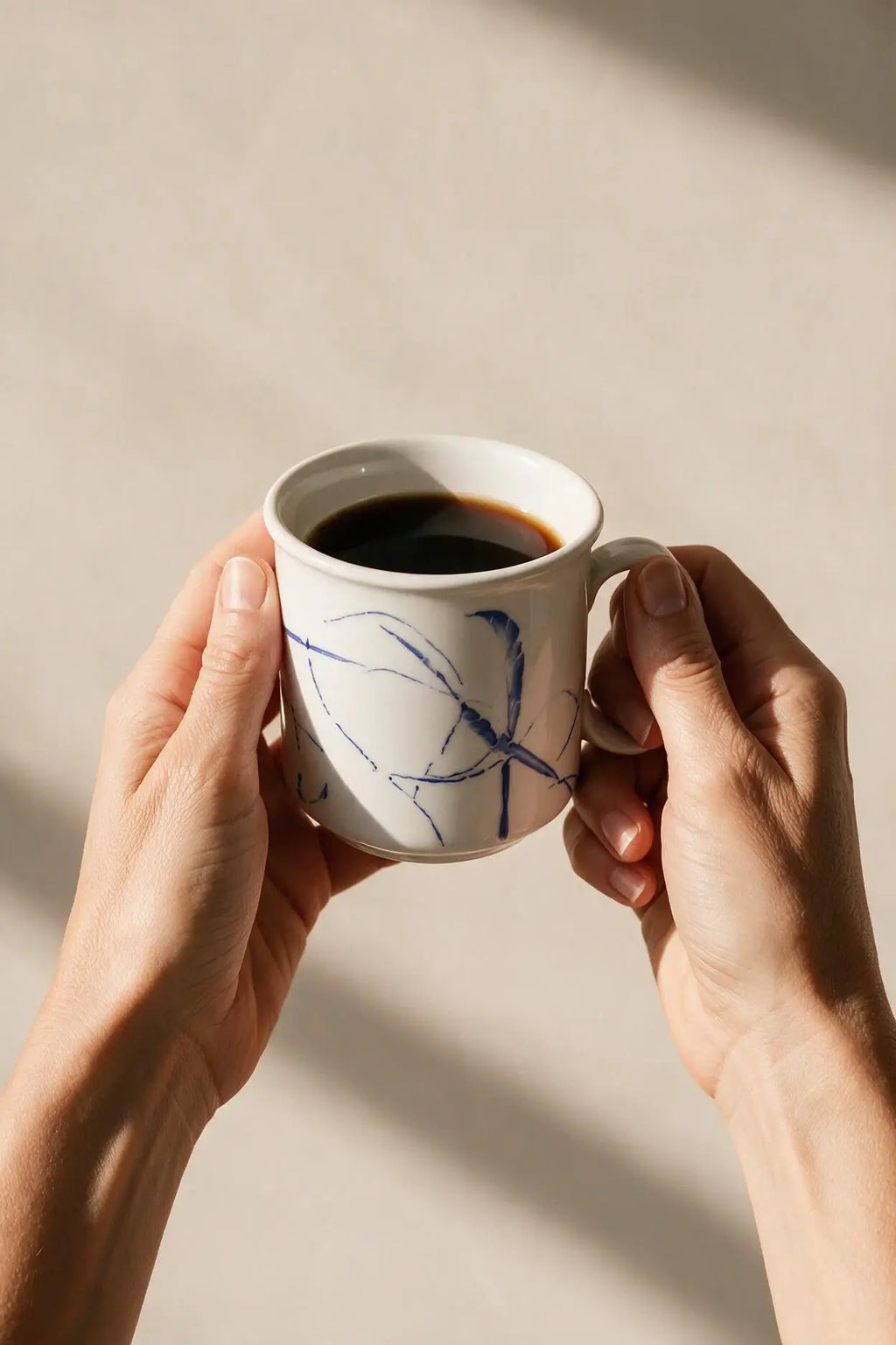 A person holds a white ceramic coffee mug with blue abstract line drawings, filled with black coffee, against a warm beige background with soft shadows.