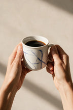 A person holds a white ceramic coffee mug with blue abstract line drawings, filled with black coffee, against a warm beige background with soft shadows.