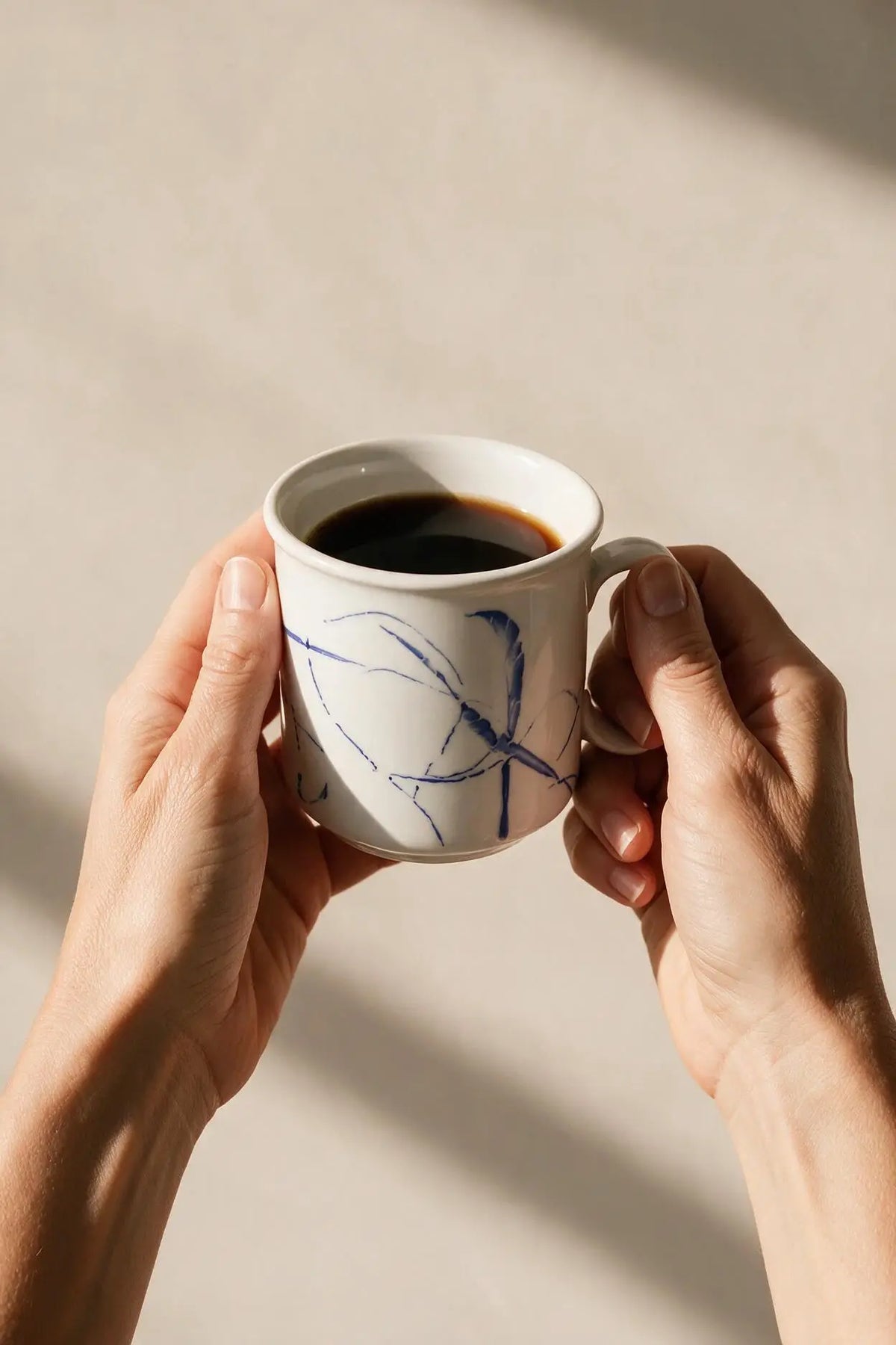 A person holds a white ceramic coffee mug with blue abstract line drawings, filled with black coffee, against a warm beige background with soft shadows.
