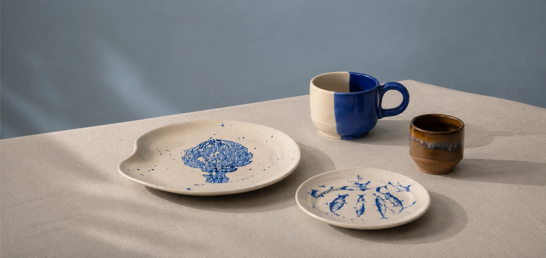 A set of ceramic tableware on a draped beige cloth against a soft blue-gray background, including a large oval plate, a small round plate, a blue-and-white mug, and a small brown cup.