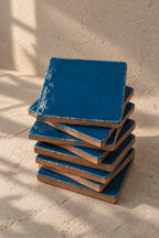 A stack of square teal blue coasters with a distressed, textured finish, photographed on a neutral beige textured background.