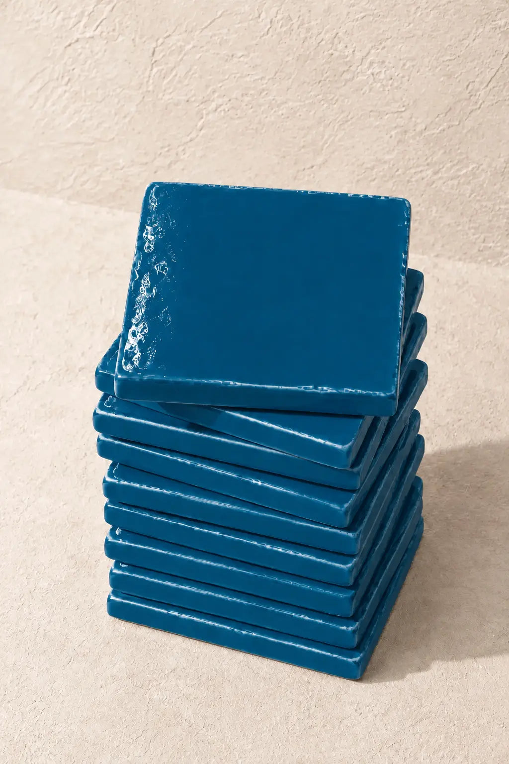 A stack of square teal blue coasters with a distressed, textured finish, photographed on a neutral beige textured background.