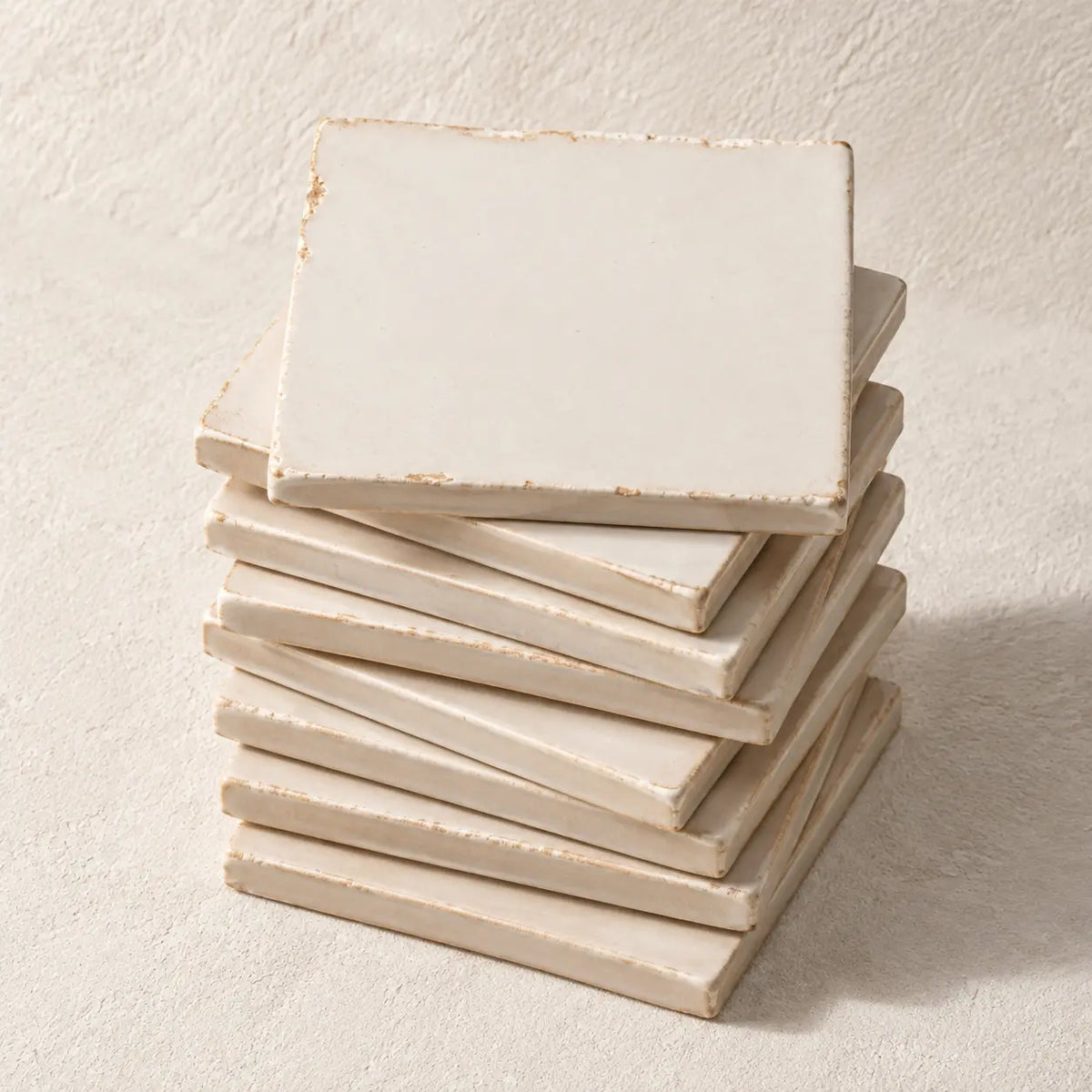 A stack of nine square ceramic coasters with a distressed, off-white finish, photographed on a textured beige background.