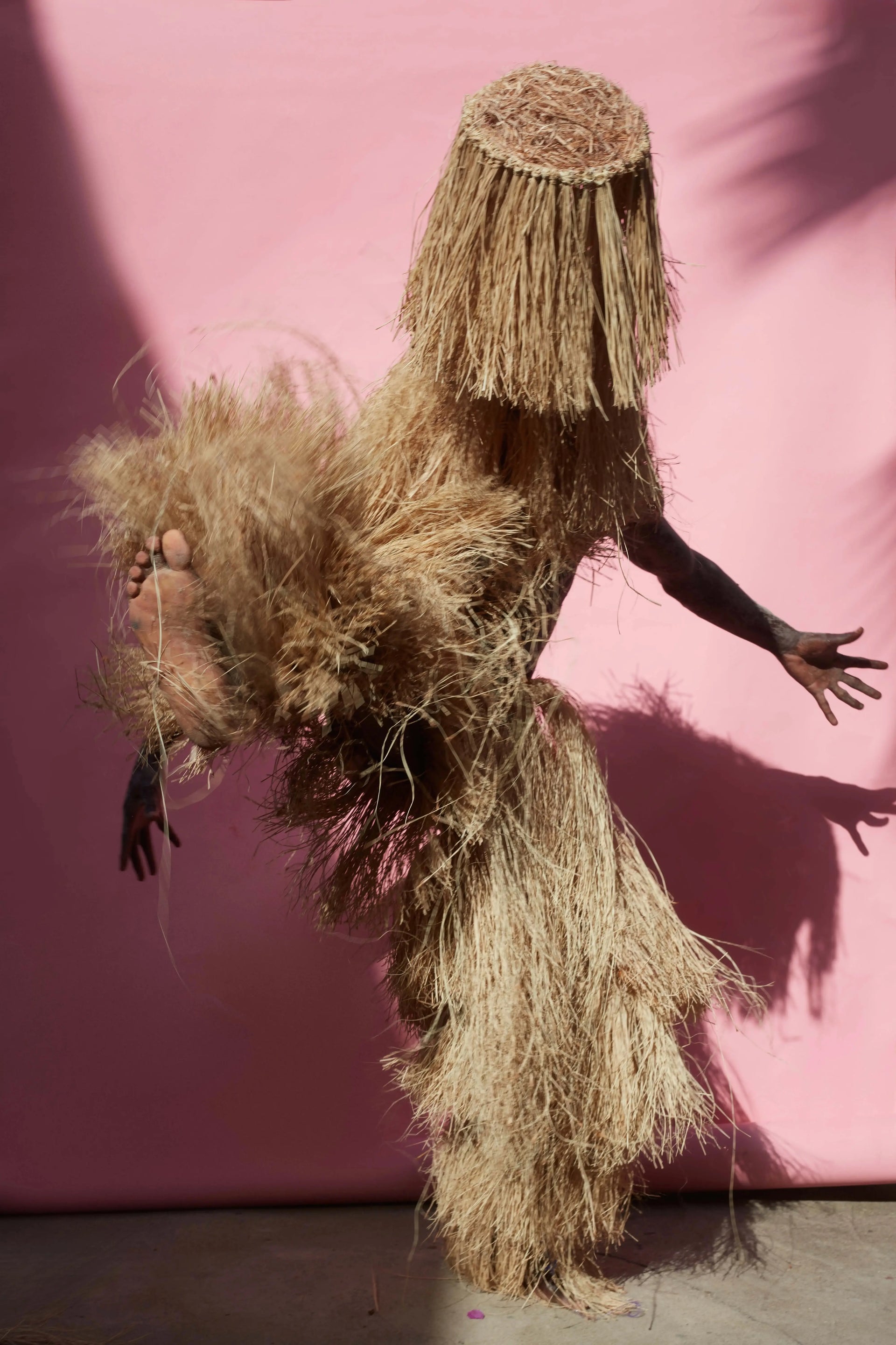A person wearing a voluminous straw or raffia costume with a long fringed head covering and dark bare arms, posed against a solid pink backdrop.