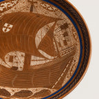 Close-up of a decorative ceramic plate or tray showing a detailed nautical scene with a large sailing ship, fish, and ornate border patterns in warm brown and cream tones with a blue band.