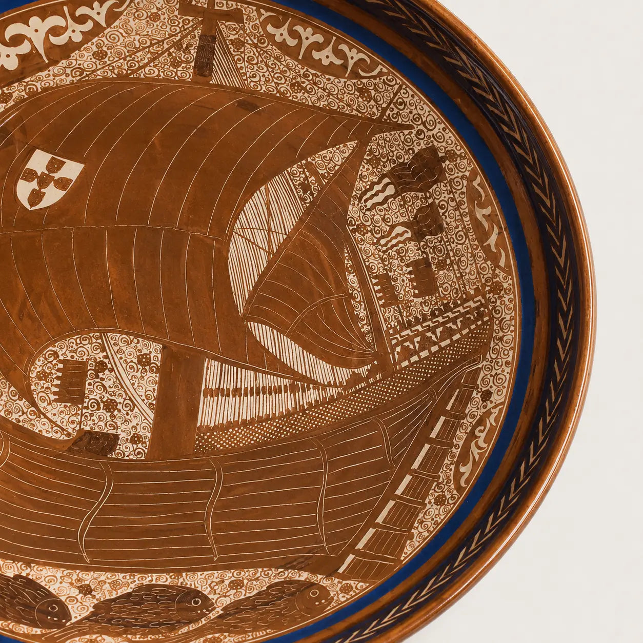 Close-up of a decorative ceramic plate or tray showing a detailed nautical scene with a large sailing ship, fish, and ornate border patterns in warm brown and cream tones with a blue band.