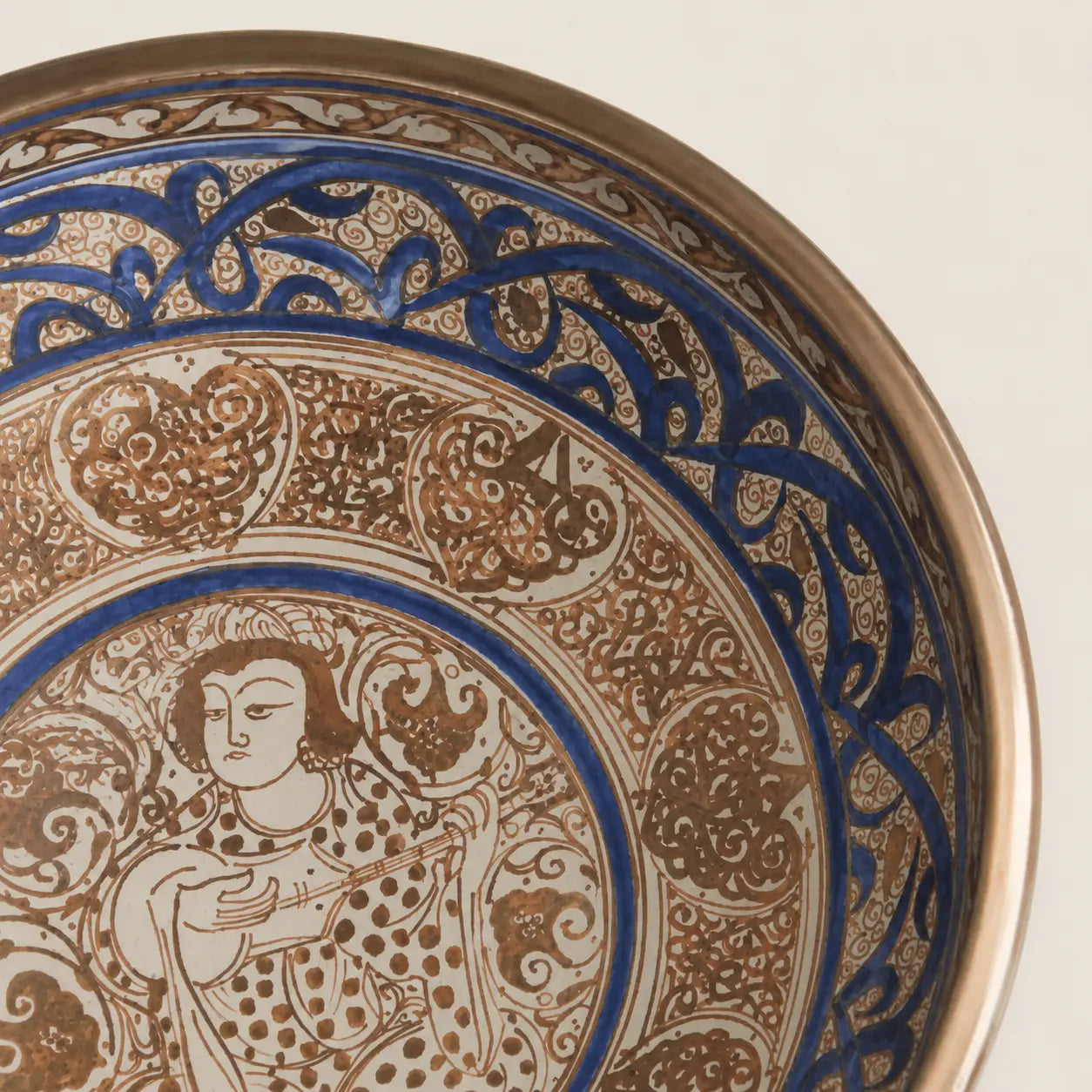 Close-up of an ornate ceramic plate or bowl interior showing a detailed hand-painted design with a figure and intricate floral scrollwork in blue, brown, and beige tones.
