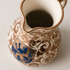 A close-up of a ceramic pitcher with a brown and white scrollwork pattern and a blue lion crest, photographed on a light neutral background.