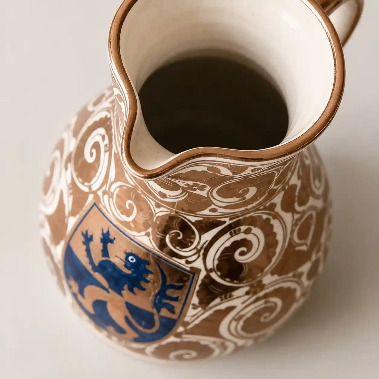 A close-up of a ceramic pitcher with a brown and white scrollwork pattern and a blue lion crest, photographed on a light neutral background.