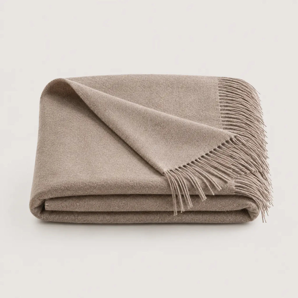 A folded beige throw blanket with fringed edges, displayed on a clean, light off-white background.