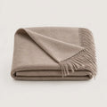A folded beige throw blanket with fringed edges, displayed on a clean, light off-white background.