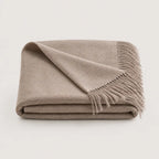 A folded beige throw blanket with fringed edges, displayed on a clean, light off-white background.