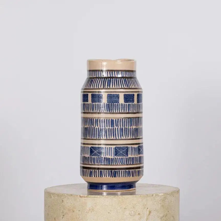 Hand-thrown ceramic vase with banded cobalt blue geometric and dash pattern on a cream tin-glaze, lustreware finish, hand-painted with silver and cobalt minerals in Spain — displayed on a stone plinth.