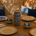 A styled dining table set with blue and yellow patterned ceramic plates and bowls, with a central blue-and-white patterned vase. The scene is set in a warm, rustic environment with a textured stone wall background and blue-and-white checkered pillows.