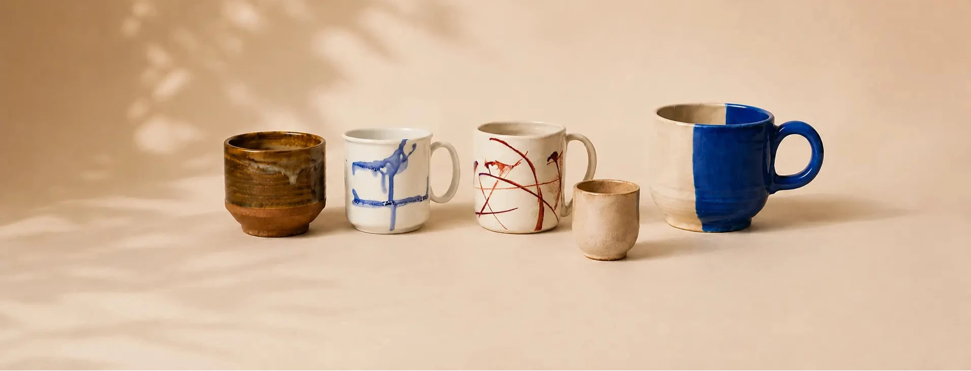Five ceramic mugs in various designs and glazes arranged on a warm beige surface with soft leaf shadows. The mugs include a brown glazed mug, a white mug with blue abstract lines, a white mug with red abstract lines, a small beige ceramic cup, and a large mug with a blue half and white half design.