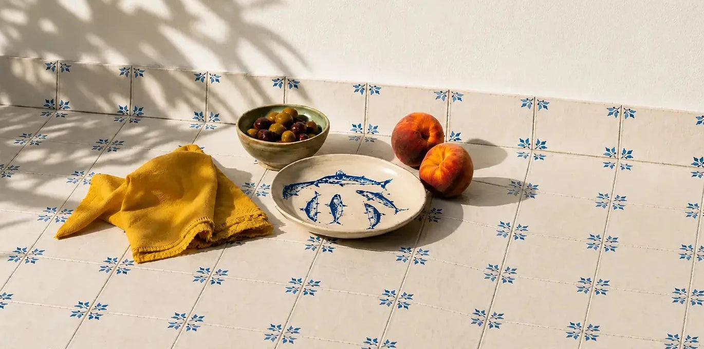 A styled tabletop scene with a white ceramic plate featuring a blue fish pattern, a folded yellow cloth, a small bowl of olives, and two oranges on a tiled surface with a white wall behind it.