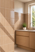 A modern kitchen corner featuring glossy vertical sand beige subway tiles on the wall, light wood cabinets, a white countertop, and a window with natural light casting diagonal shadows.