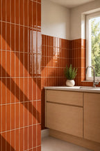 Orange glossy rectangular wall tiles in terracotta orange colour, installed in a kitchen corner with a light wood cabinet, white countertop, and a small potted green plant near a window.